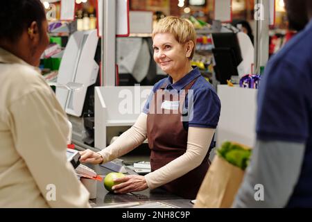 Felice bionda maturo cassiere guardando il consumatore afro-americano femmina mentre il costo di conteggio della mela verde matura dal banco di cassa Foto Stock