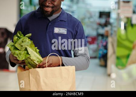 Primo piano di un giovane assistente di vendita maschile afro-americano in uniforme blu che mette le verdure nel sacchetto di carta mentre aiuta i clienti Foto Stock