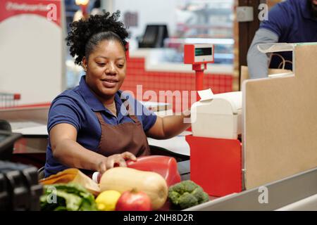 Giovane assistente del negozio afro-americano in uniforme prendendo i prodotti alimentari dalla linea di checkout prima di scannerizzare il loro prezzo Foto Stock