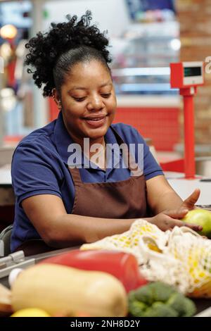 Felice giovane donna cassiere in uniforme seduta presso il banco di cassa in supermercato e scansione prodotti alimentari dei clienti Foto Stock