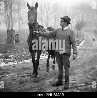 1965, storico, invernale e all'aperto su un sentiero fangoso, un uomo con un cavallo, si è adornato per un giro invernale mattutino, Inghilterra, Regno Unito. Il manager della stalla o forse l'allenatore, indossa una camicia e cravatta, un maglione, un cappello e un paio di bretelle o un paio di jodhpurs e stivali. Foto Stock