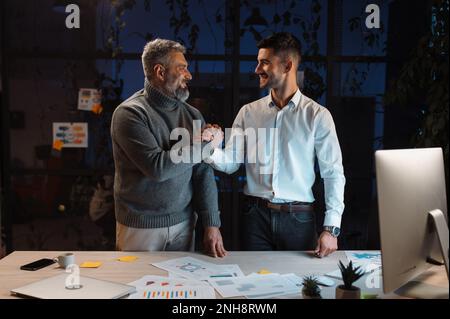 Il manager maschile stringe la mano a un collega senior esperto in ufficio serale Foto Stock