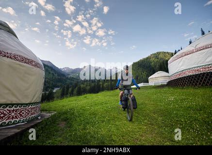 Uomo con greggi e barba in mountain bike con borse turistiche vicino a Nomad Yurt casa nella bella valle di montagna in Almaty, Kazakistan. Estrem Foto Stock