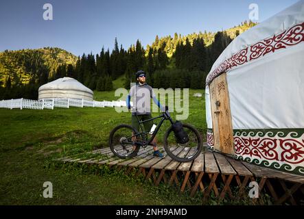 Uomo con greggi e barba in piedi con mountain bike con borse turistiche vicino a casa Nomad Yurt nella bella valle di montagna in Almaty, Kazakistan. Foto Stock