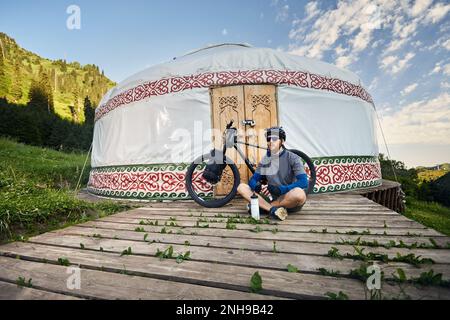 Uomo con greggi e barba seduto di fronte alla mountain bike con borse turistiche vicino a casa Nomad Yurt nella bella valle di montagna in Almaty, kazako Foto Stock