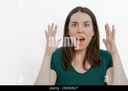 Bruna donna dai capelli lunghi con bocca aperta sorpresa guardando la macchina fotografica con le braccia sollevate sfondo bianco Foto Stock