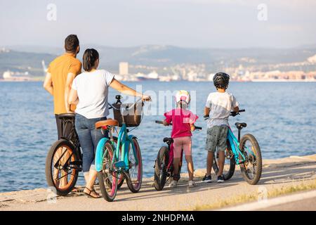 Famiglia caucasica con due bambini in piedi in bicicletta e godersi il tramonto sulla riva del mare, vista posteriore. Foto Stock