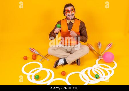 Ritratto di un giocoliere in un giubbotto e trucco seduto con gli oggetti su uno sfondo giallo Foto Stock