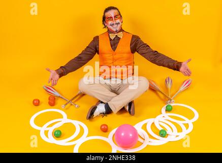 Ritratto di un giocoliere in un giubbotto e trucco seduto con gli oggetti di giocoleria su uno sfondo giallo Foto Stock