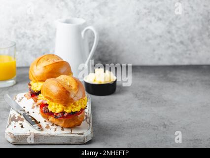 Sandwich per la colazione con uova strapazzate, pancetta, formaggio, pomodoro su tavola di legno bianco, bicchiere con succo d'arancia fresco, sfondo bianco. Fare Foto Stock