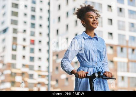 Ragazza felice adolescente afroamericana in abito blu casual guardando in avanti mentre si guida scooter elettrico contro edifici moderni Foto Stock