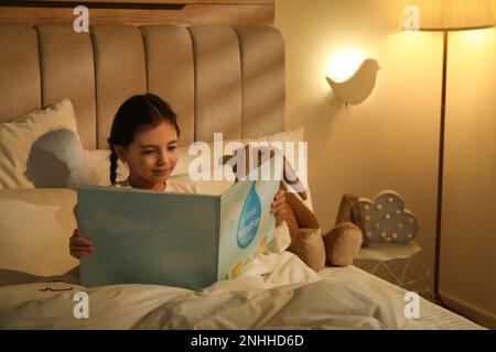 Piccola ragazza lettura libro in camera da letto illuminato da lampada da notte Foto Stock