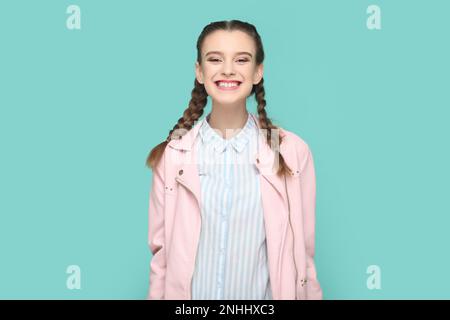 Ritratto di estremamente felice ragazza giovane allegra con trecce indossando giacca rosa in piedi guardando la macchina fotografica con un sorriso toothy, sorride ampiamente. Studio in interni isolato su sfondo verde. Foto Stock