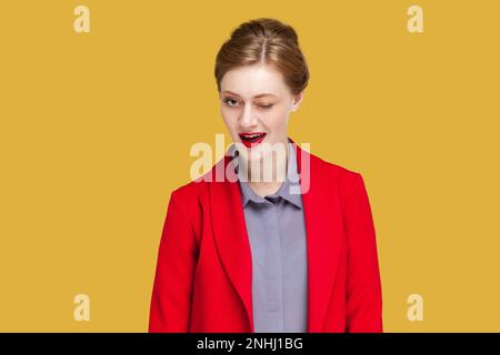 Ritratto di donna flirting positivo con labbra rosse che le lampeggiano gli occhi con piacere avendo espressione felice, indossando giacca rossa. Studio al coperto isolato su sfondo giallo. Foto Stock