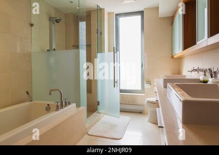 Lavandino con ripiano in granito con i suoi lavandini, vasca da bagno bianca con rivestimento in marmo, box doccia in vetro nel bagno con pavimento in piastrelle di marmo all'interno dell'appartamento. Foto Stock