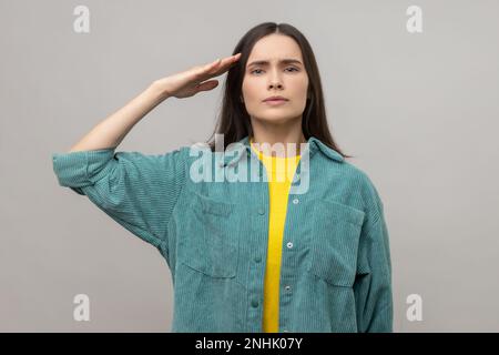 Sì Signore. Giovane donna seria e intelligente che dà saluto, ascoltando con attenzione l'ordine, guardando con responsabile espressione attenta, indossando giacca casual. Studio in interni isolato su sfondo grigio. Foto Stock