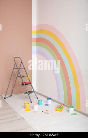 Diversi utensili decoratori e scala vicino alla parete con arcobaleno dipinto al coperto Foto Stock