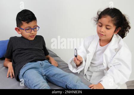 Latino ragazza e ragazzo giocare per essere un medico con un cappotto bianco che è troppo grande per lei e paziente che agisce per sentirsi malato molto divertente Foto Stock