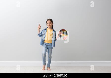 Bambina con pennello e tavolozza di vernice vicino alla parete grigio chiaro al coperto Foto Stock