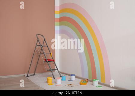 Diversi utensili decoratori e scala vicino alla parete con arcobaleno dipinto al coperto Foto Stock