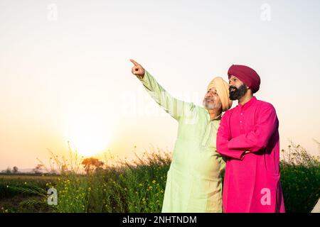 Ritratto di felice punjabi srdar padre e figlio con kurta e pagdi turbano in piedi e puntando qualcosa a distanza in campo agricolo. Copia Foto Stock