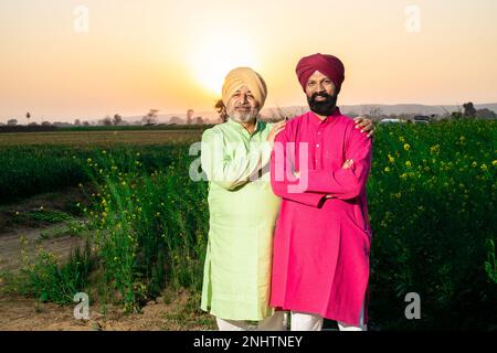 Ritratto di felice padre e figlio punjabi che indossano kurta e pagdi turbano in piedi insieme in campo agricolo. Foto Stock