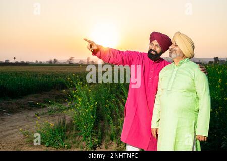 Ritratto di felice punjabi srdar padre e figlio con kurta e pagdi turbano in piedi e puntando qualcosa a distanza in campo agricolo. Copia Foto Stock