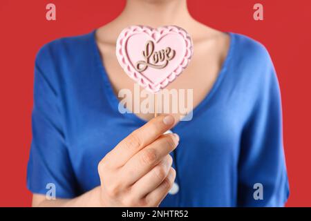 Donna che tiene lollipop a forma di cuore fatto di cioccolato su sfondo rosso, primo piano Foto Stock