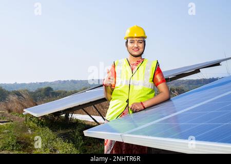 Ritratto di giovane donna indiana tecnico indossando un elmetto giallo in piedi vicino a pannelli solari. Industrial lavoratore impianto solare, alternativa Foto Stock