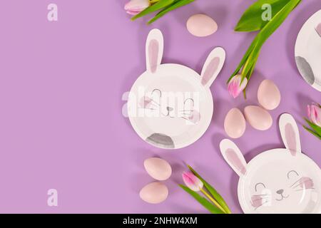 Festa di Pasqua con piatto di carta a forma di coniglietto, uova e fiori di tulipano su sfondo viola con spazio copia Foto Stock