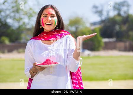 Ritratto di felice giovane donna indiana indossando kurta bianco e dupatta vestito tenuta polvere colore lastra fare gesto mano per la pubblicità celebrando h Foto Stock