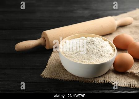 Ciotola di farina, uova e rolling pin su tavola di legno nero Foto Stock