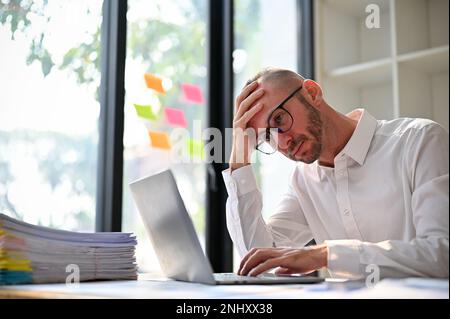 Sottolineato e premuroso uomo d'affari caucasico o lavoratore di ufficio maschio che hanno alcuni problemi sul suo progetto, lavorando sul suo progetto online sul laptop in th Foto Stock