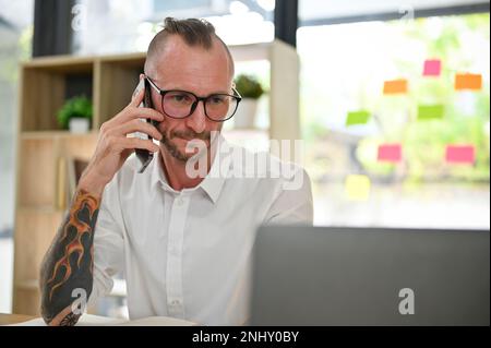 Uomo d'affari caucasico intelligente o imprenditore maschile con occhiali e tatuaggio sulle sue braccia è al telefono con un potenziale cliente, chiamando a qualcuno mentre Foto Stock