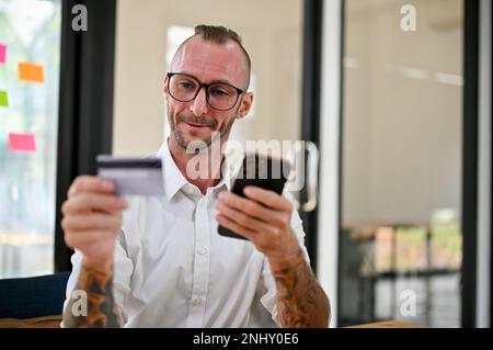 Felice uomo caucasico con gli occhiali e tatuaggio sulle braccia si siede alla sua scrivania tenendo una carta di credito o di debito e smartphone, utilizzando il mobile banking per p Foto Stock