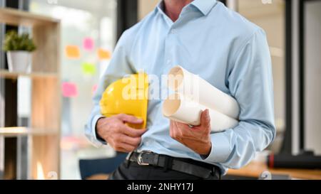 Immagine ritagliata di un ingegnere o architetto professionista che tiene un elmetto giallo e dei progetti in ufficio. Foto Stock