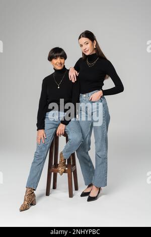 intera lunghezza di donna elegante in piedi con mano in tasca di jeans vicino felice madre asiatica seduta su sgabello di legno su sfondo grigio Foto Stock