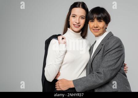 madre e figlia asiatica in abiti eleganti abbracciando e guardando la macchina fotografica isolata sul grigio Foto Stock