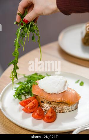 Una ragazza mette la rucola su un piatto con un panino e un uovo in camicia. Concetto di colazione. Foto Stock