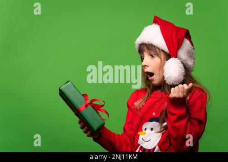 Ragazza emozionale in un maglione lavorato a maglia e cappello di Babbo Natale sorprende un regalo fantastico. Indovina la sorpresa. Buon Natale e Felice anno nuovo 2023. Foto di alta qualità Foto Stock
