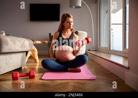 Donna incinta che indossa l'abbigliamento di idoneità sul tappeto a casa esercitandosi con i pesi Foto Stock