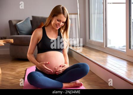 Donna incinta che indossa l'abbigliamento di idoneità sul tappeto di esercitazione a casa che fa lo yoga Foto Stock