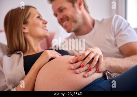 Coppia amorevole a casa sdraiata sul letto con l'uomo che tocca lo stomaco della donna incinta Foto Stock