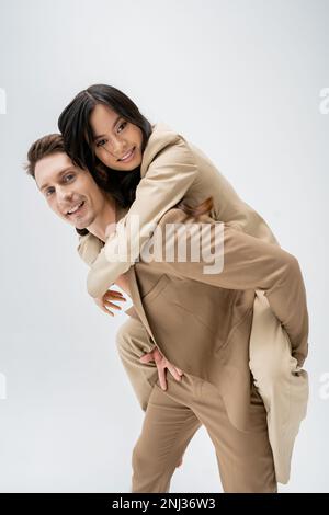 felice ed elegante uomo piggybacking giovane donna asiatica, mentre sorridendo alla fotocamera isolato su grigio, immagine stock Foto Stock
