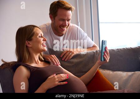 Coppia con donna incinta Holding Mobile Phone relax su divano a casa Holding stomaco Foto Stock