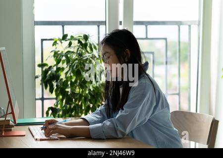 Studente asiatico concentrato ragazza in eyewear impianti home computer su progetto di ricerca formazione online. Foto Stock