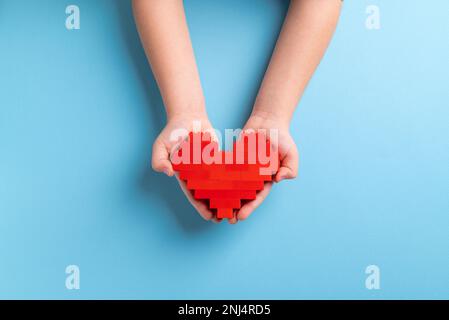 Mani di bambina che tiene il cuore fatto di mattoni di plastica su sfondo blu Foto Stock