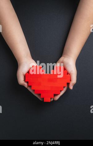 Mani di bambina che tiene il cuore fatto di mattoni di plastica su sfondo grigio scuro Foto Stock