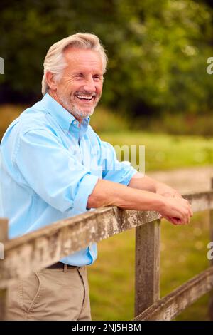 Ritratto di sorridente uomo maturo o anziano vestito casualmente Pendente su Fence a piedi in campagna Foto Stock