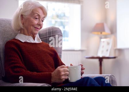 Donna anziana con bevanda calda seduta in poltrona nella Lounge at Home Foto Stock